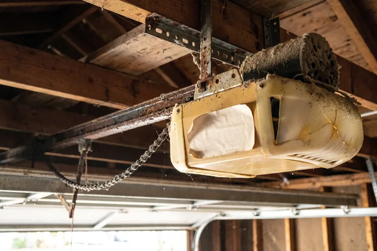 An old chain-drive garage door opener on a garage ceiling, its housing yellowed and dusty and the chain sagging